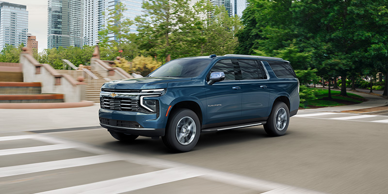 2026 Chevrolet Suburban stopped at a crosswalk on a lovely green-tree-lined city center street.