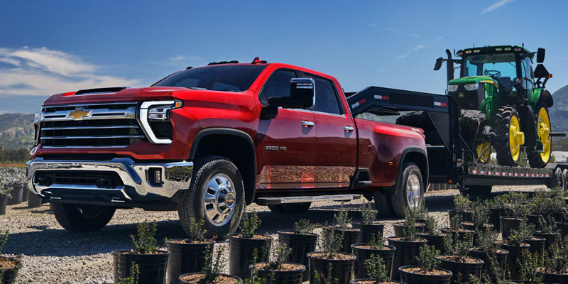 A red 2026 Chevrolet Silverado 3500 is hauling a large green and yellow tractor on a dirt lane at a seedling tree farm.