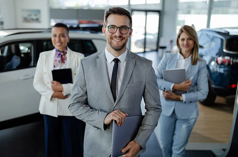 A GMC sales team smile at us in the showroom - a suited man in the front and 2 lovely suited women on e4ither side begind him = all 3 with their Ipads in hand.